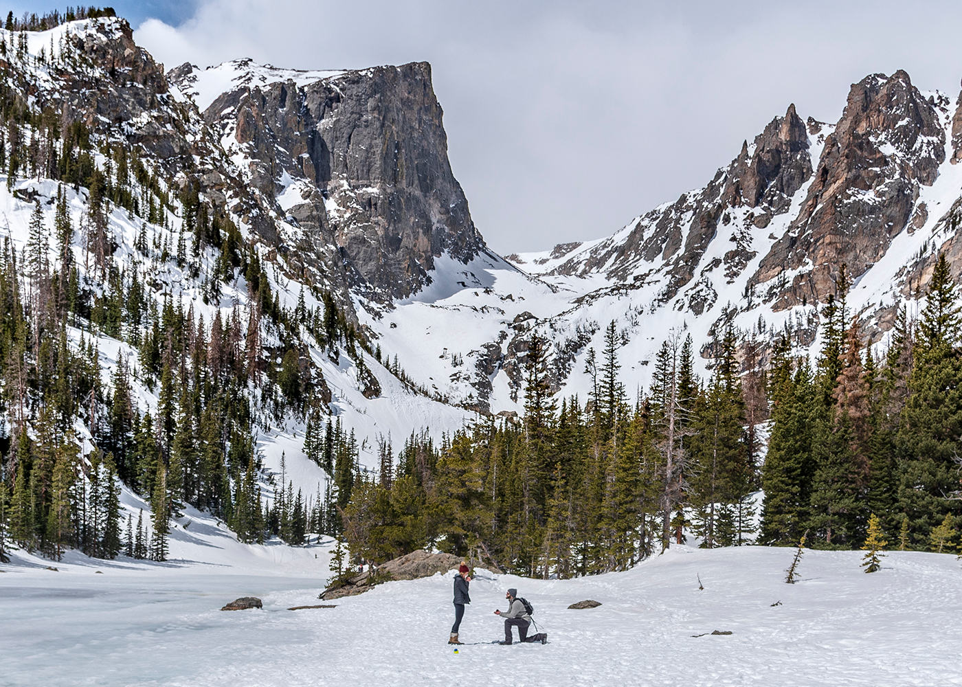 A snow-covered mountain landscape at Rocky Mountain National Park with evergreen trees while a man is down on one knee proposing marriage