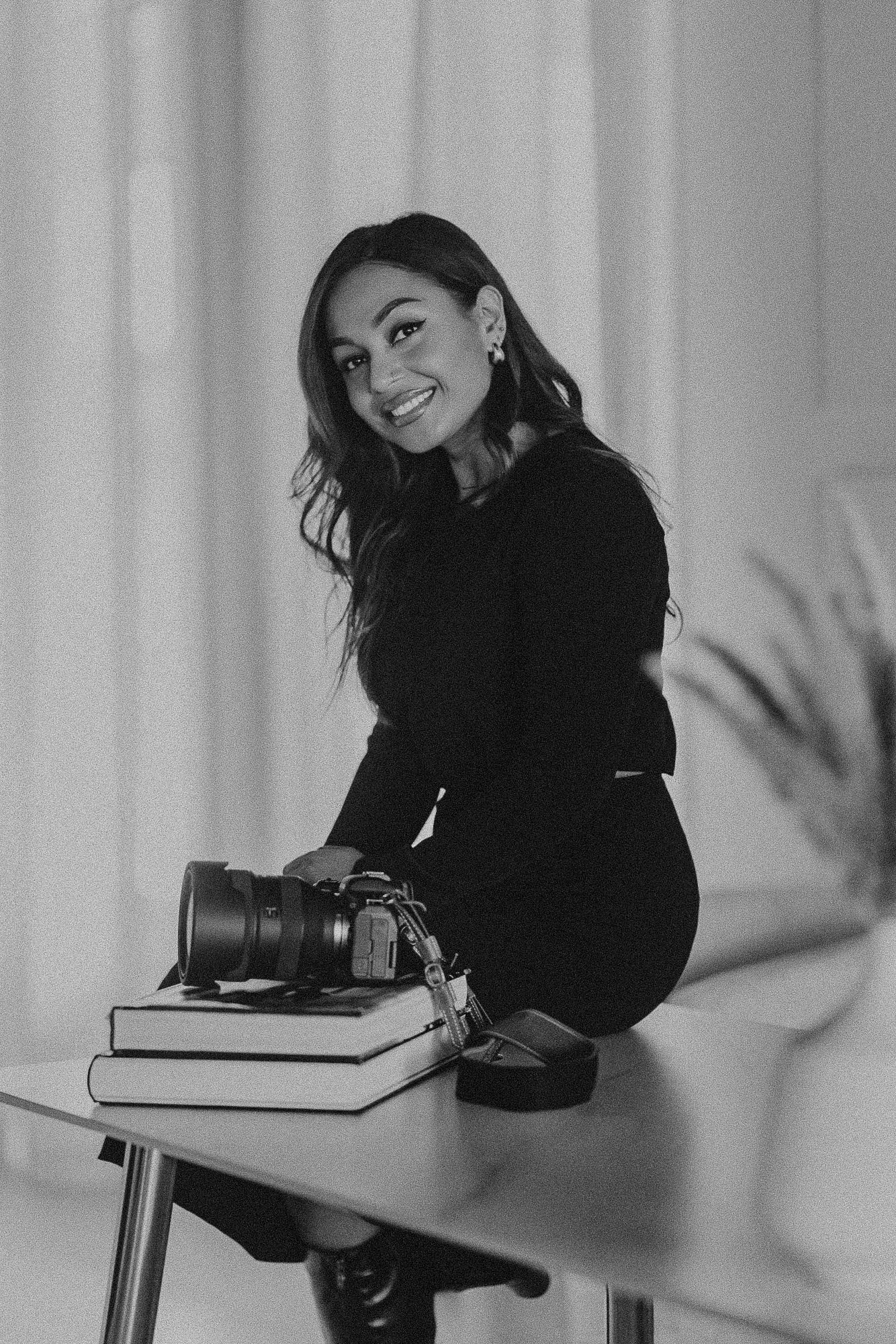 Black and white portrait of photographer in dark clothing sitting on table with camera and books in studio setting.