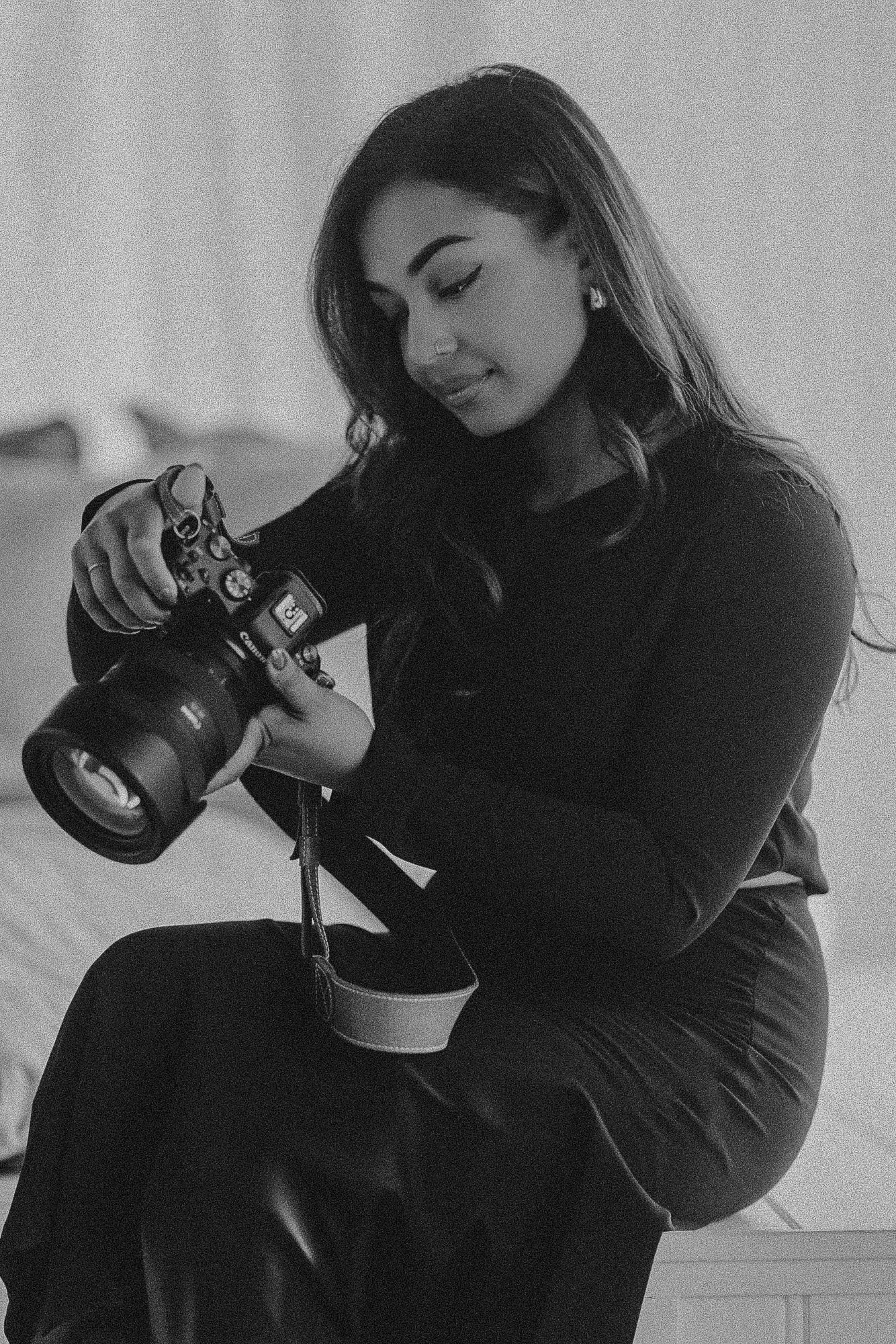 A photographer in black attire examines settings on a professional DSLR camera in a monochrome portrait.