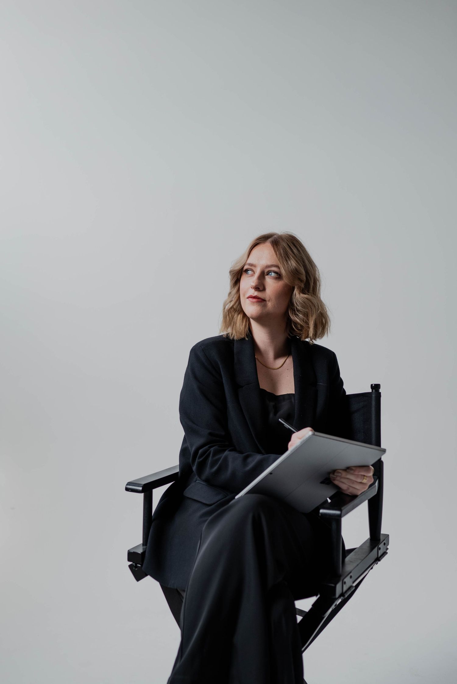 Professional in black outfit sits in director's chair reviewing documents against white background.