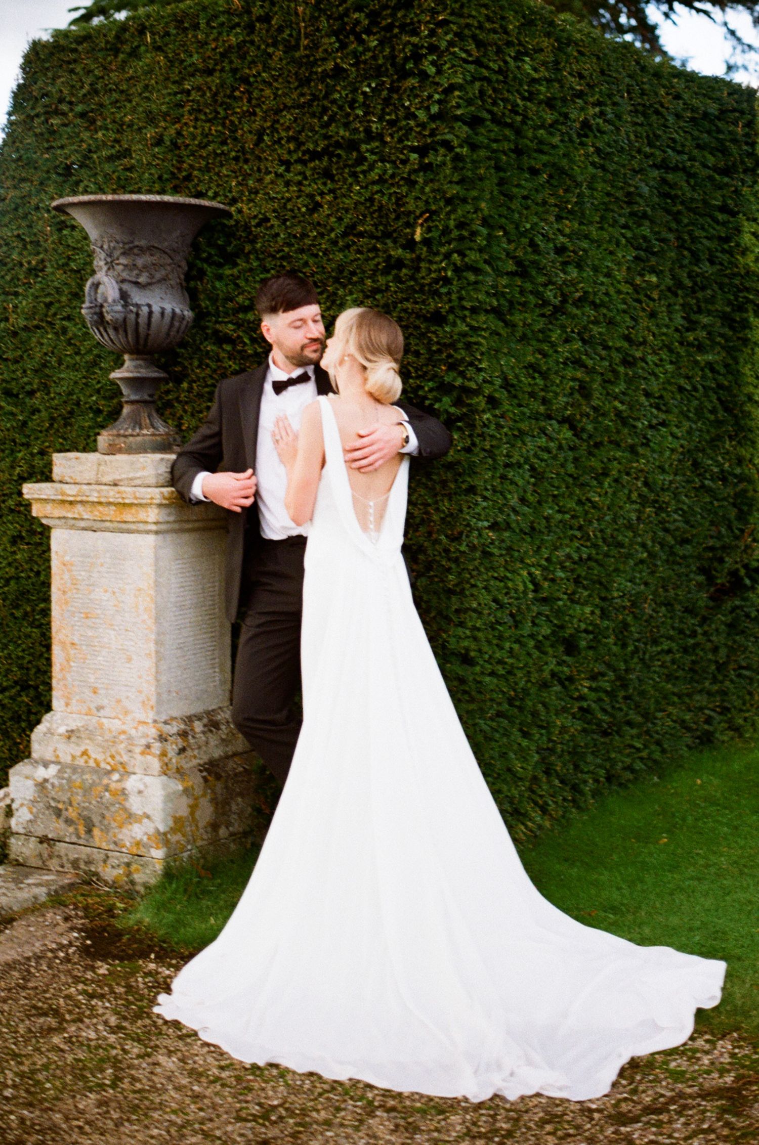 A bride and groom embrace by an ornate stone pedestal with decorative urn against a manicured hedge wall.
