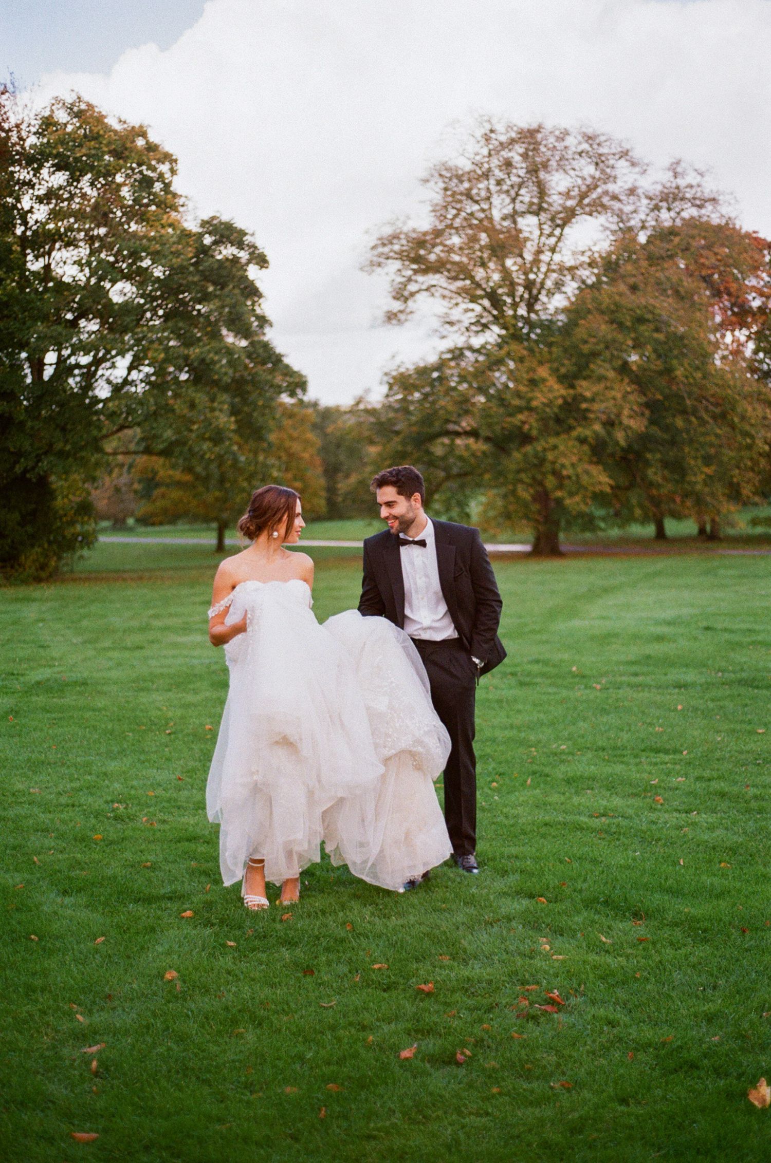 A wedding couple walks on lush green grass with autumn trees in the background at a scenic outdoor venue.
