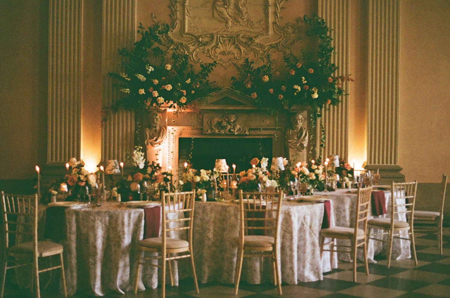 Elegant reception hall with round tables, white linens, gold chairs, and dramatic floral arrangements illuminated by candlelight.