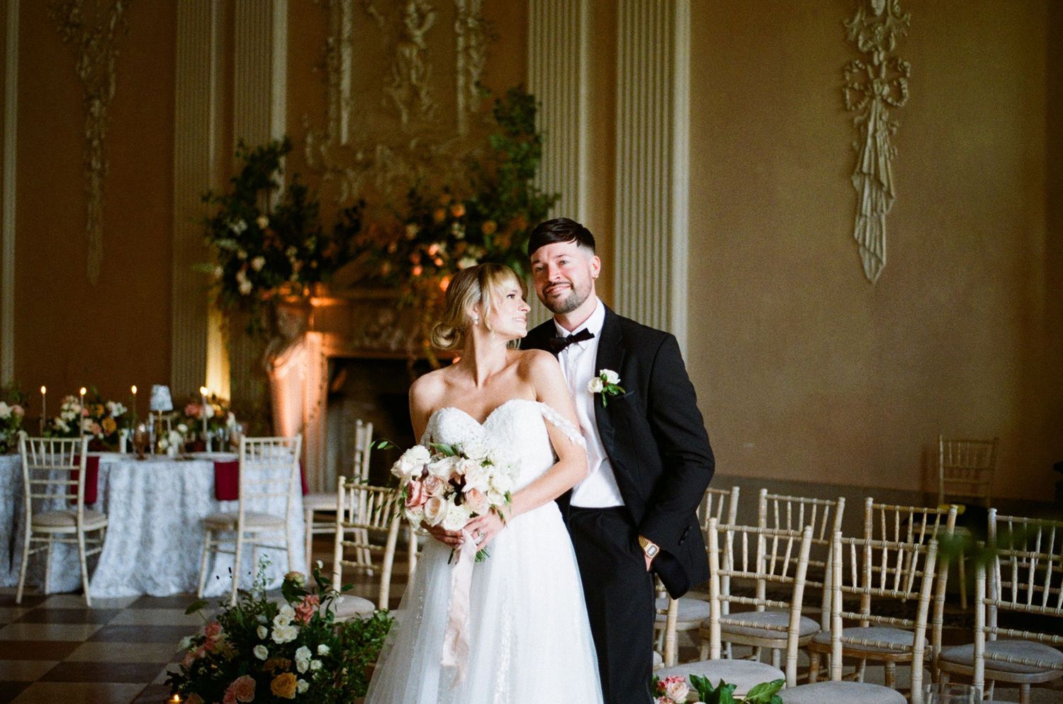 Elegant wedding reception venue decorated with floral arrangements and gold chiavari chairs in a grand ballroom setting.