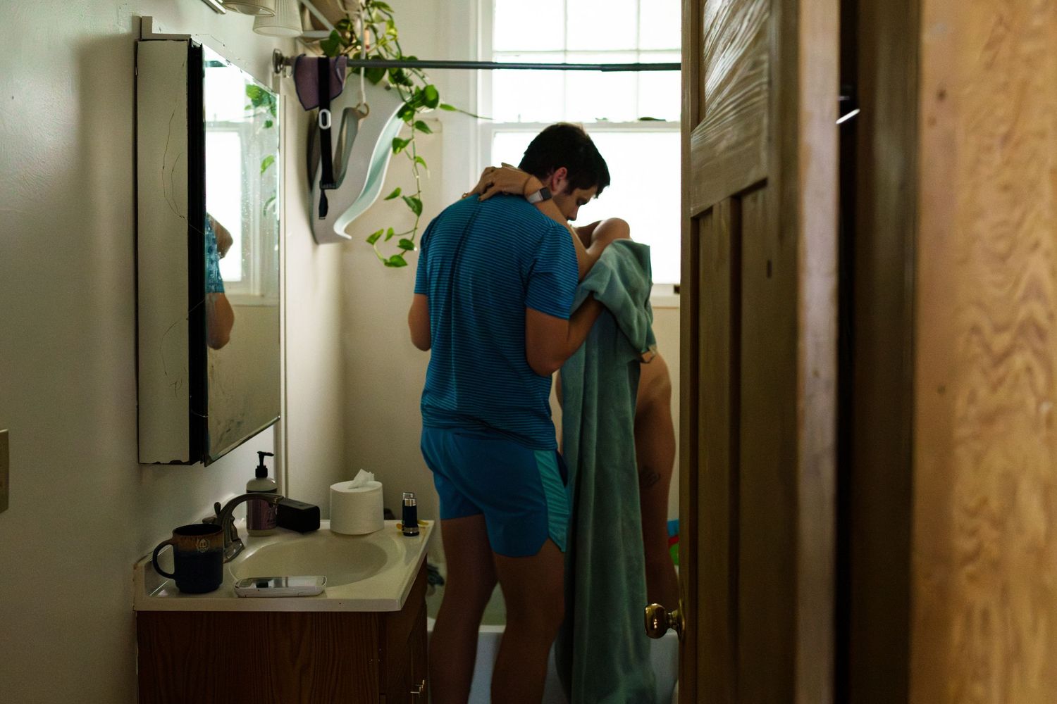 Person in teal shirt arranging houseplants in bright room near sunny window.