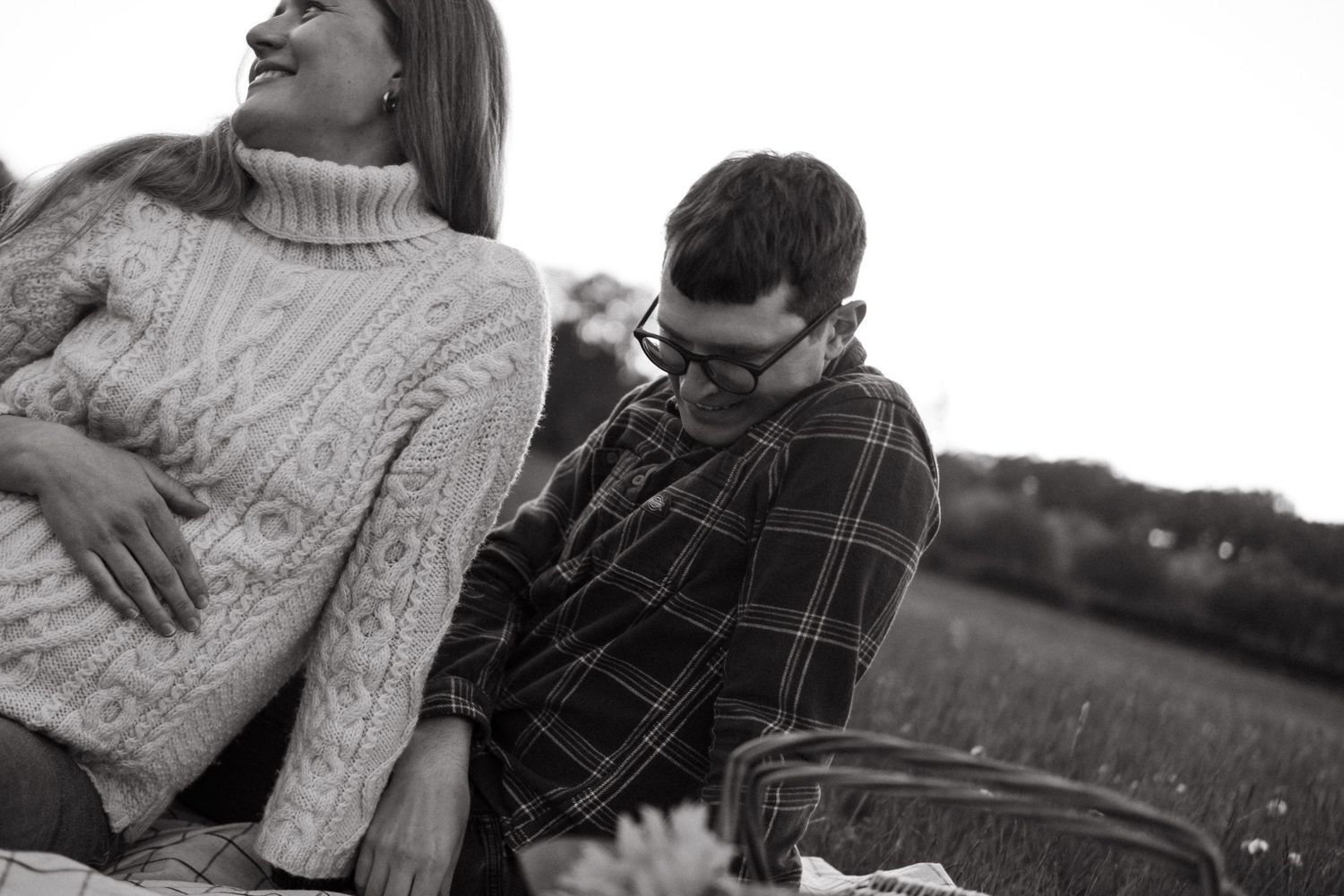 smiling couple sitting on a blanket with woman touching her pregnant belly while being captured by maternity photographer in Stockholm