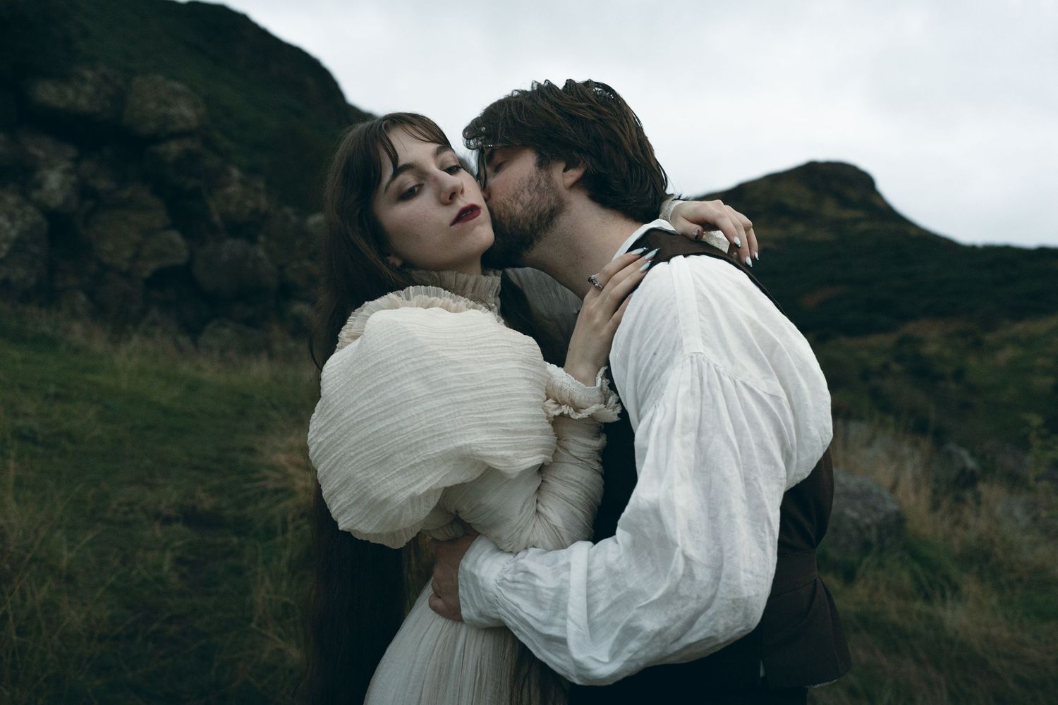 A romantic couple in flowing white attire shares intimate moments against a dramatic mountainous landscape.