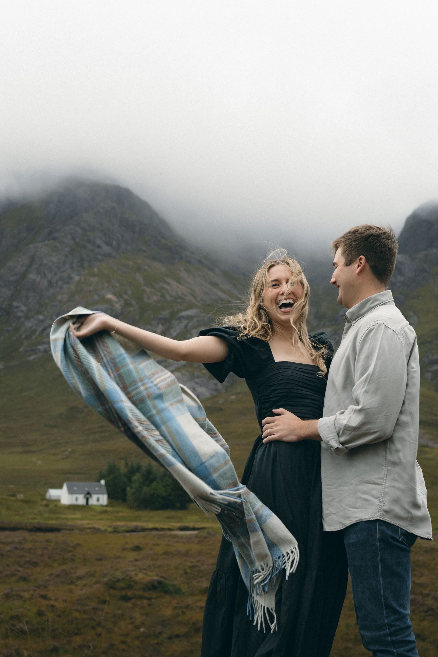 A couple shares a joyful moment in front of misty Scottish highlands with a small white house in the background.