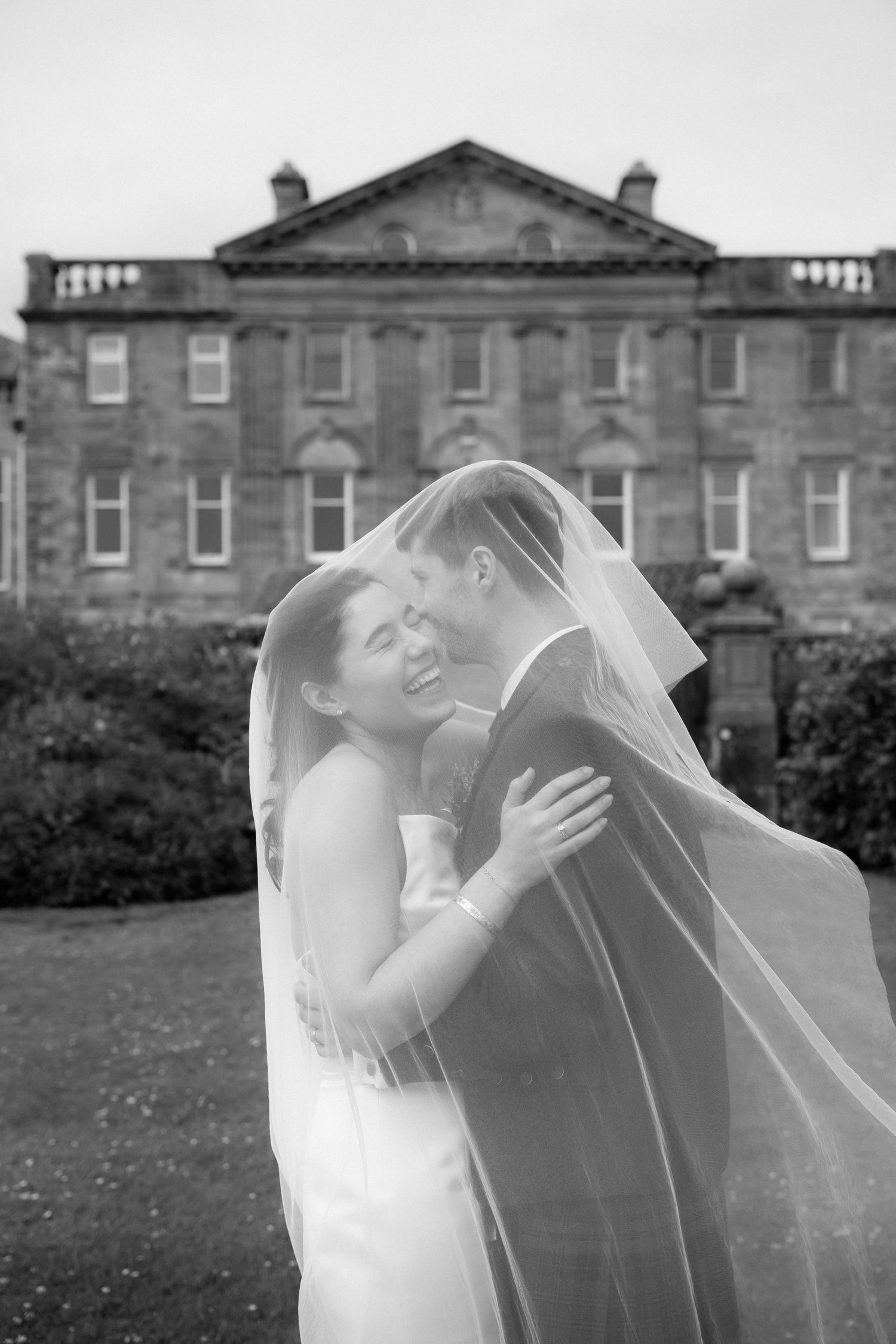 A romantic wedding moment captured in black and white outside a grand historic manor house.