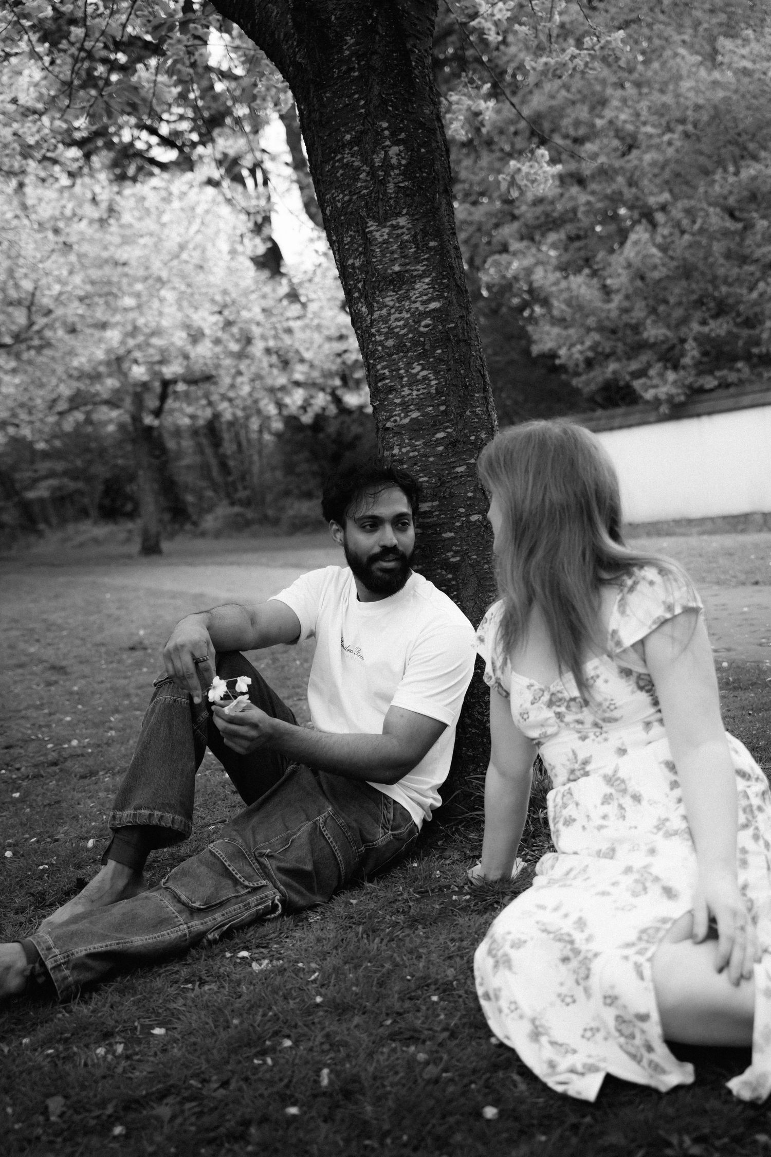 A couple sits together under a tree in a park, with one person wearing a white t-shirt and the other in a floral dress.