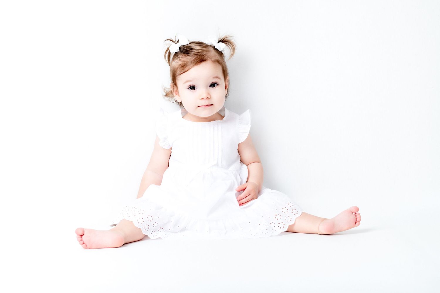 A small baby wearing a white dress sits on a white background with their legs spread out and hair accessory.
