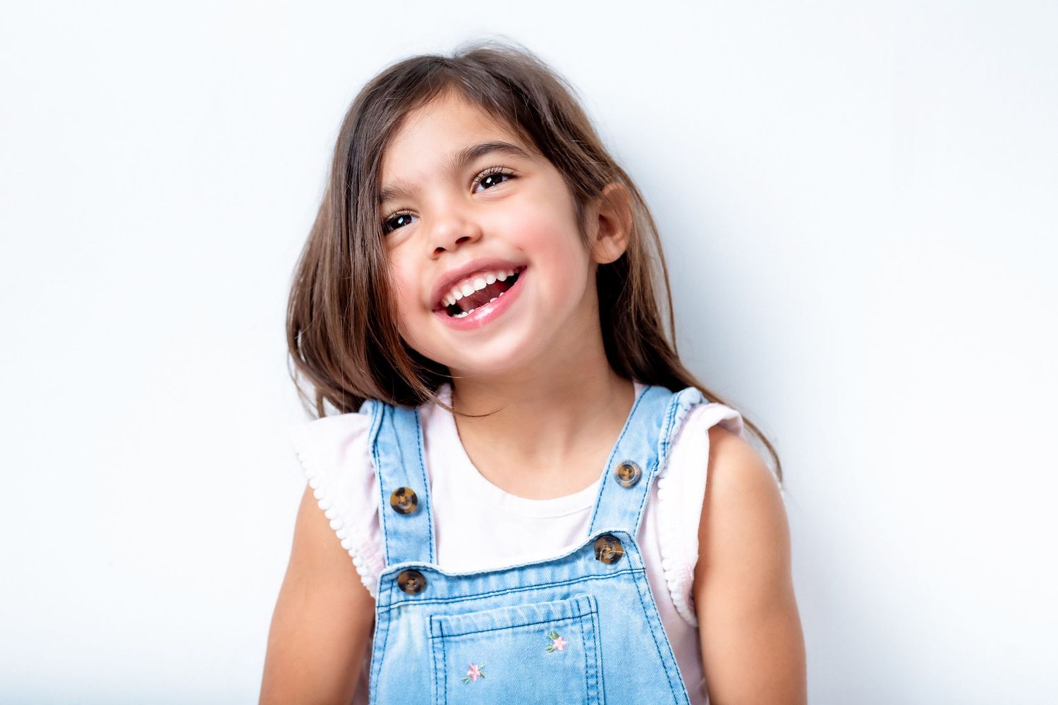A child wearing denim overalls and a white sleeveless top smiles brightly against a plain white background.