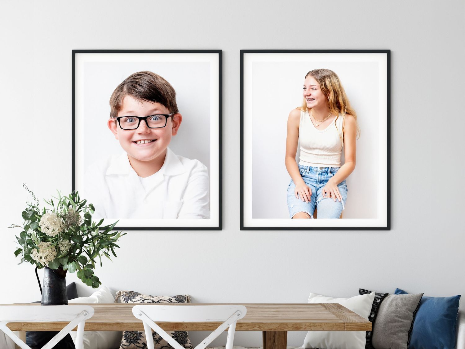 Two black-framed portrait photographs hanging on a gray wall above a white dining table with decorative plant.