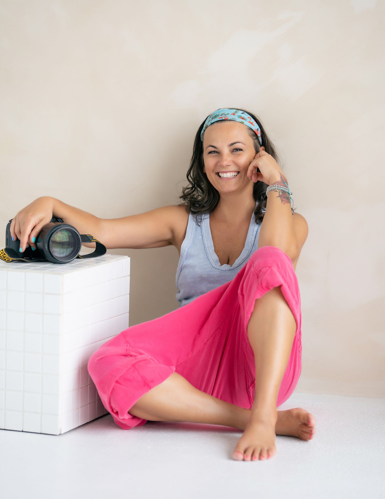 Professional photographer sitting on floor in casual blue tank top and pink pants with camera resting on white cube beside them.