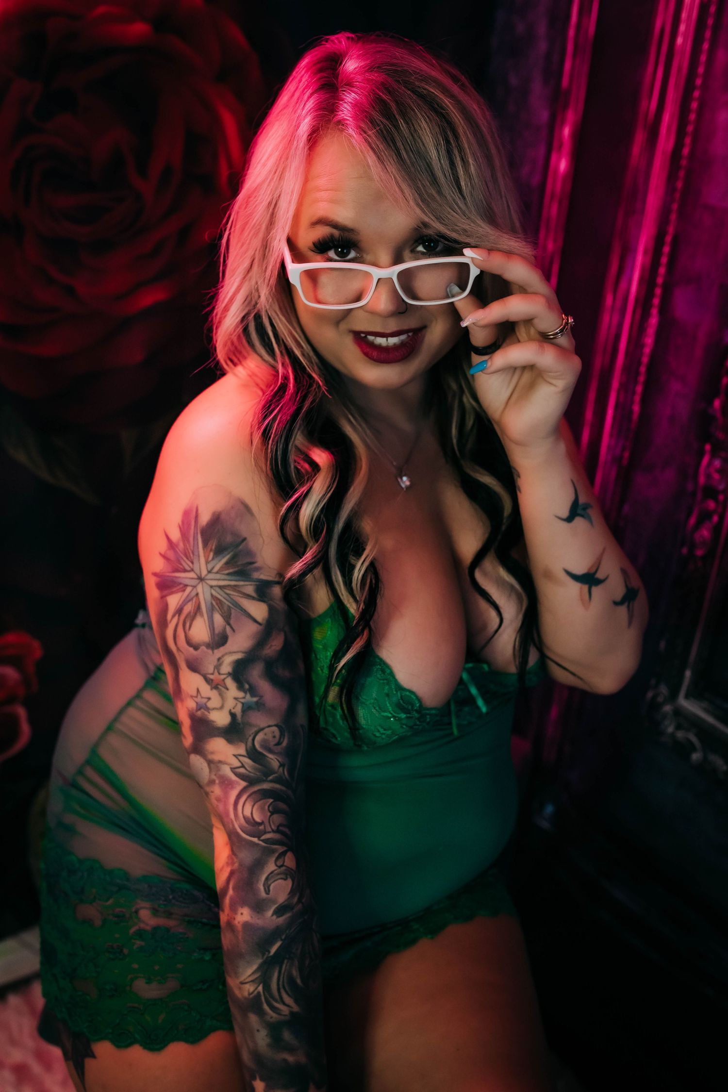 Figure in green lingerie with sleeve tattoo and glasses poses in dramatic red lighting against dark background.