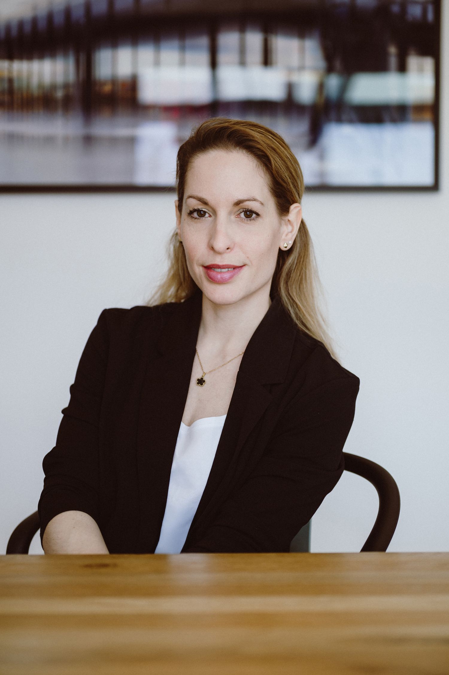 Professional business portrait of someone in a black blazer and white top seated at a wooden desk in an office setting.