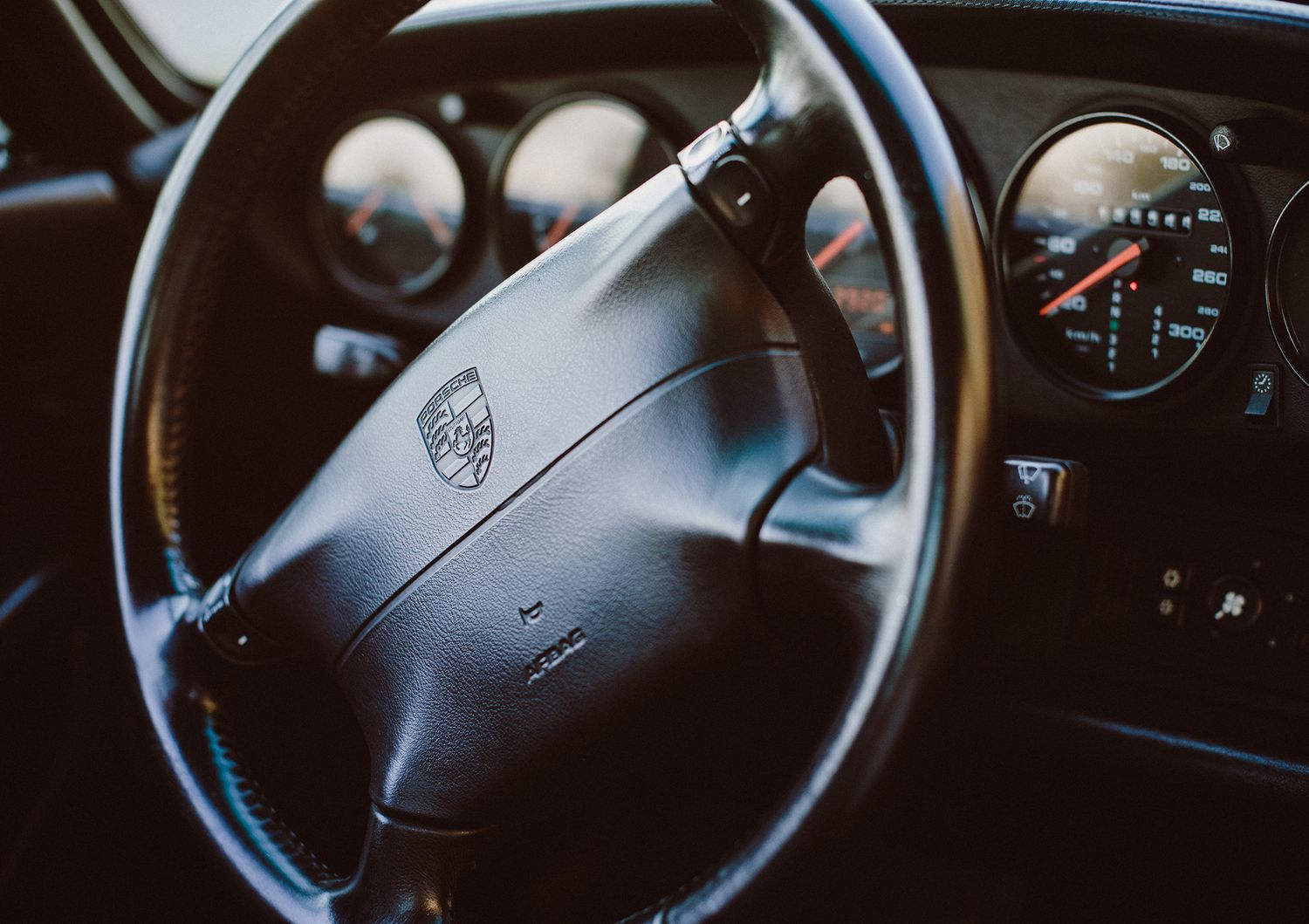 Black leather steering wheel and dashboard gauges of a Porsche automobile interior captured in dark lighting conditions.