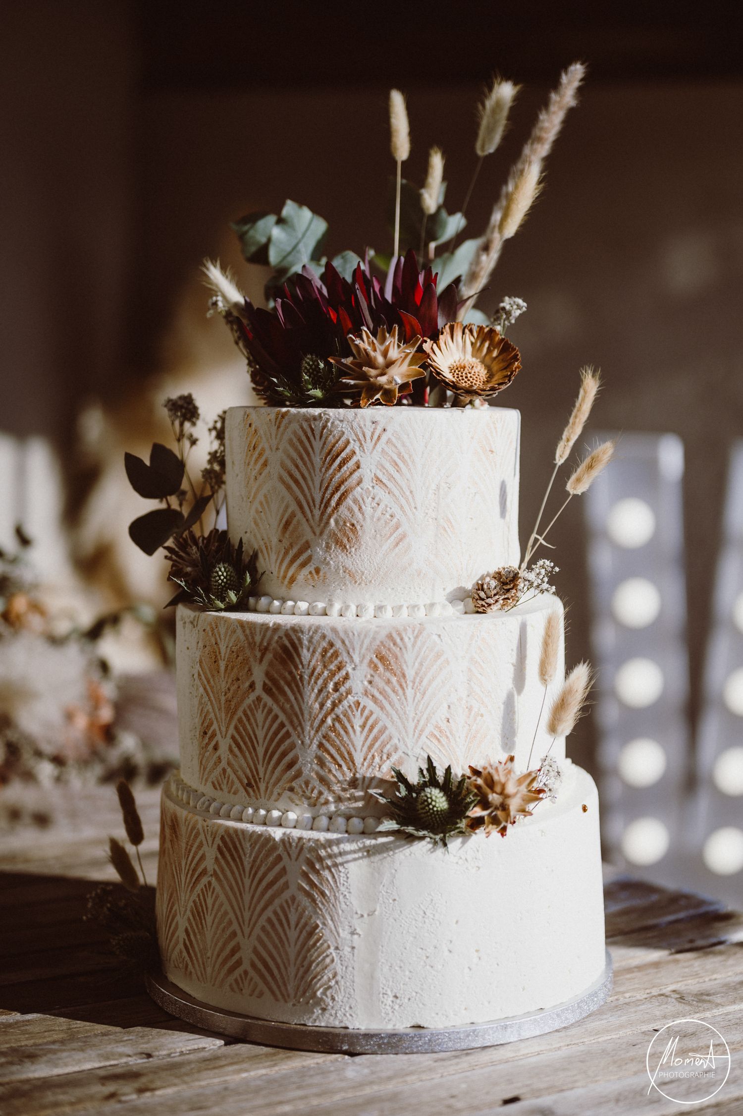 Elegant three-tier white wedding cake decorated with art deco patterns and dried flowers on rustic wooden table.
