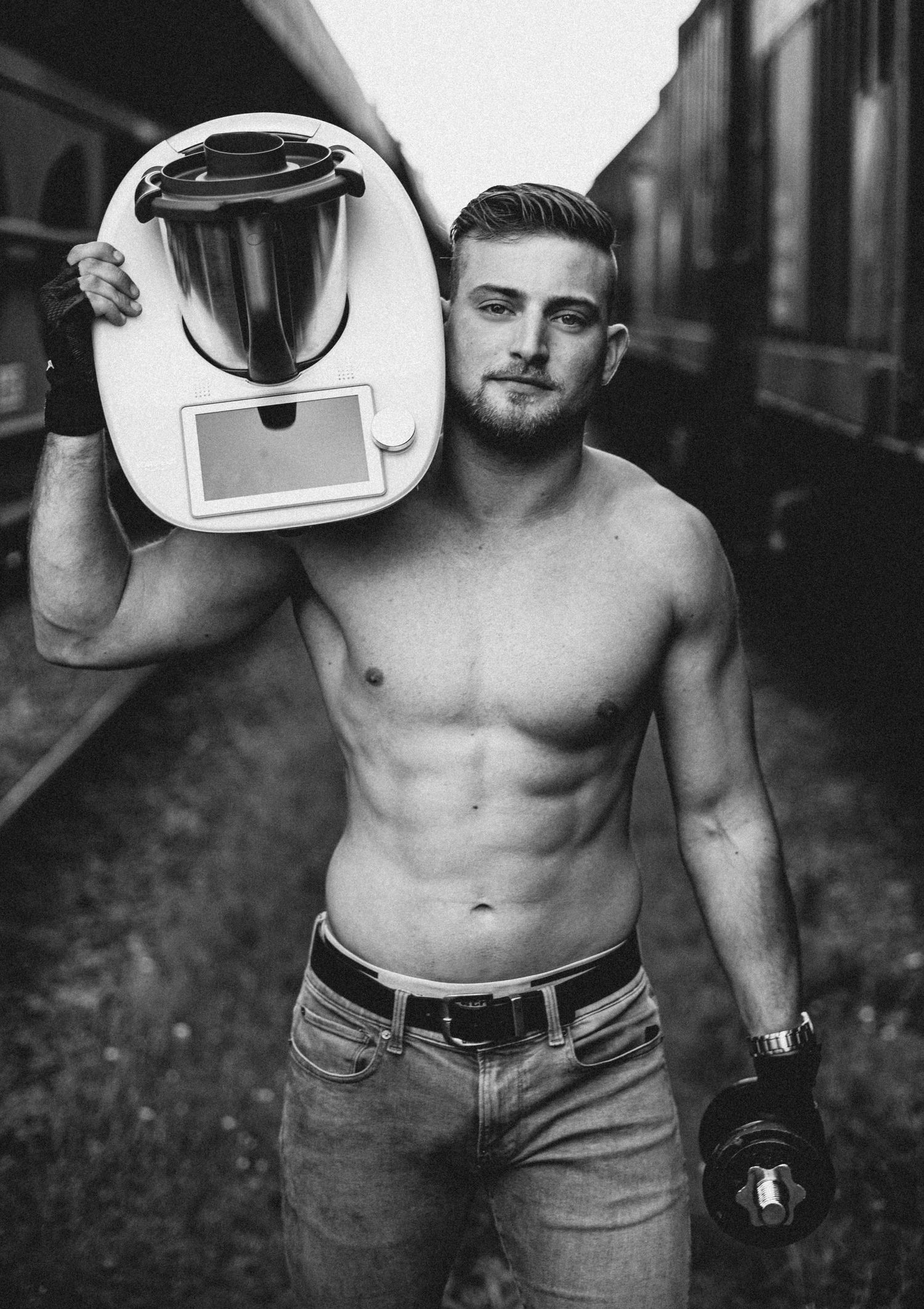 Black and white photo showing a shirtless person holding a white Thermomix appliance against an urban background.