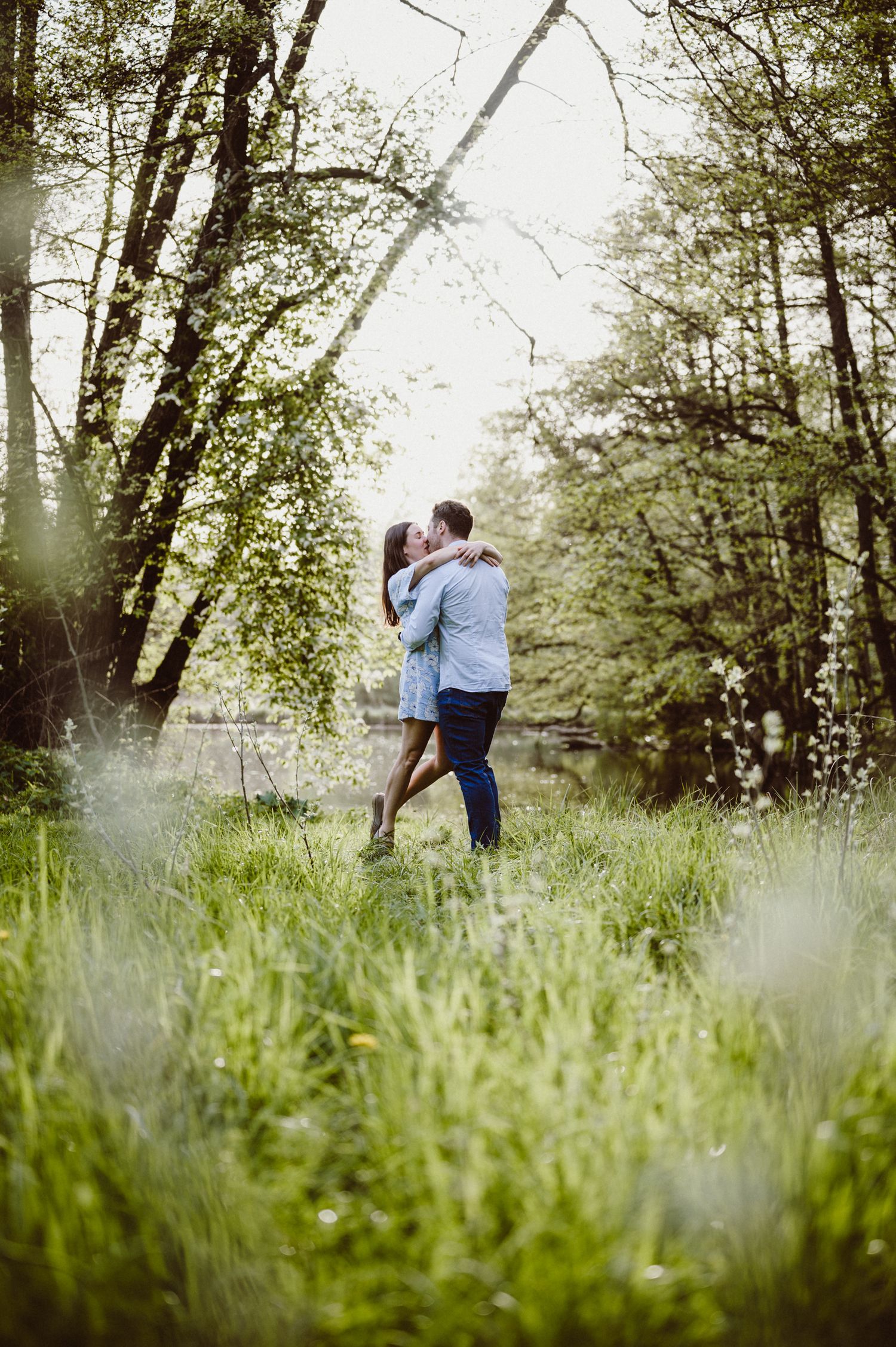 A romantic embrace captured in tall grass and sunlit woods, with soft light filtering through the trees.