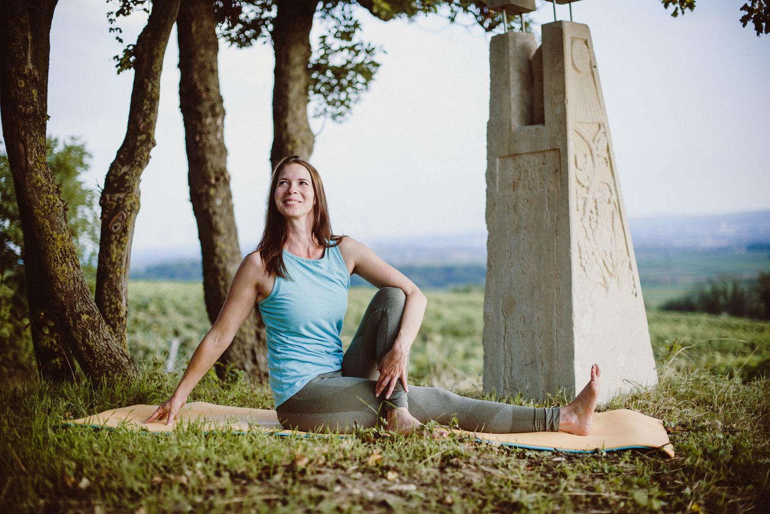 Person in blue tank top practicing yoga pose near stone monument in grassy hilltop setting.