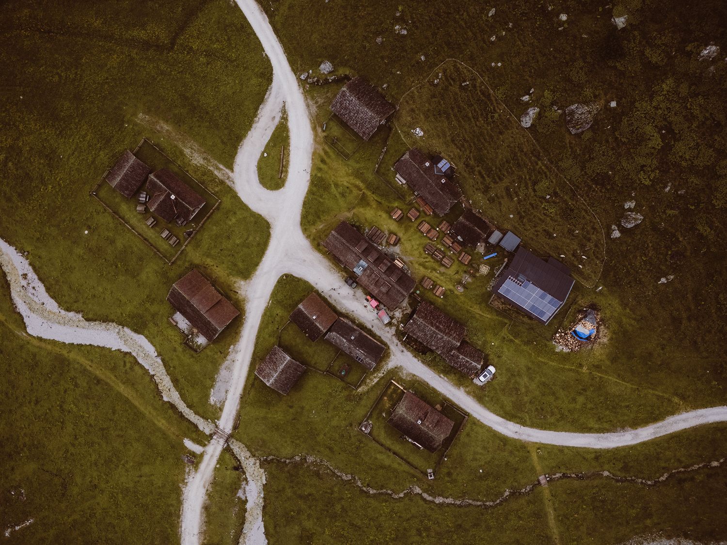 Aerial view of small mountain village with traditional wooden houses connected by winding dirt paths on green meadow.