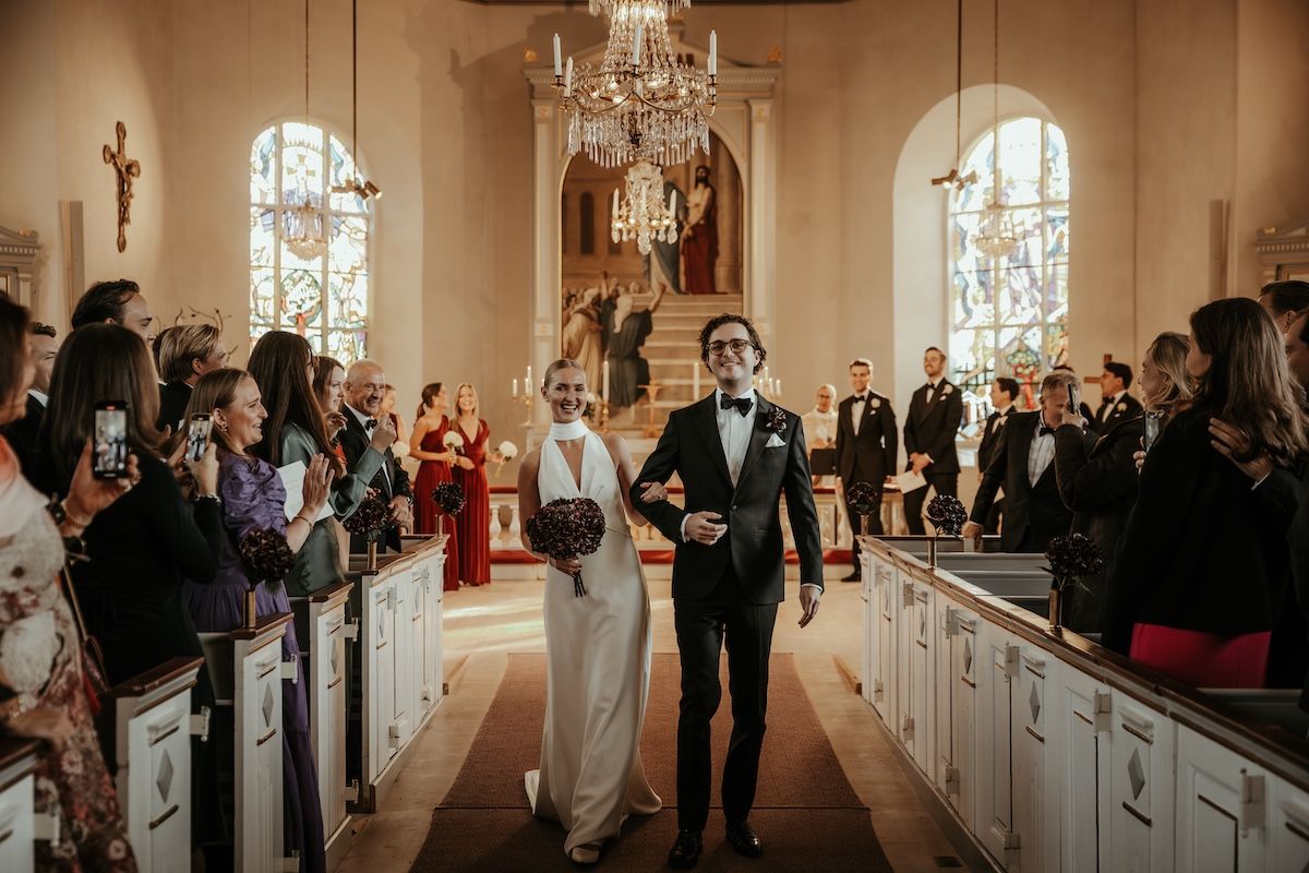 Ett brudpar går glada ner för altargången och tar emot gästernas applåder efter en vigsel i Släps kyrka. Foto av Grevholm Photography