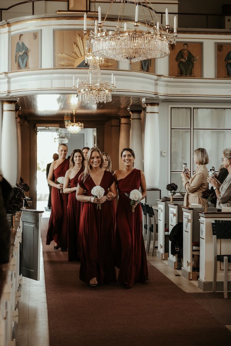 Brudtärnor iklädda röda klänningar går ner för altaret inför en kyrklig vigsel. Foto av Grevholm Photography