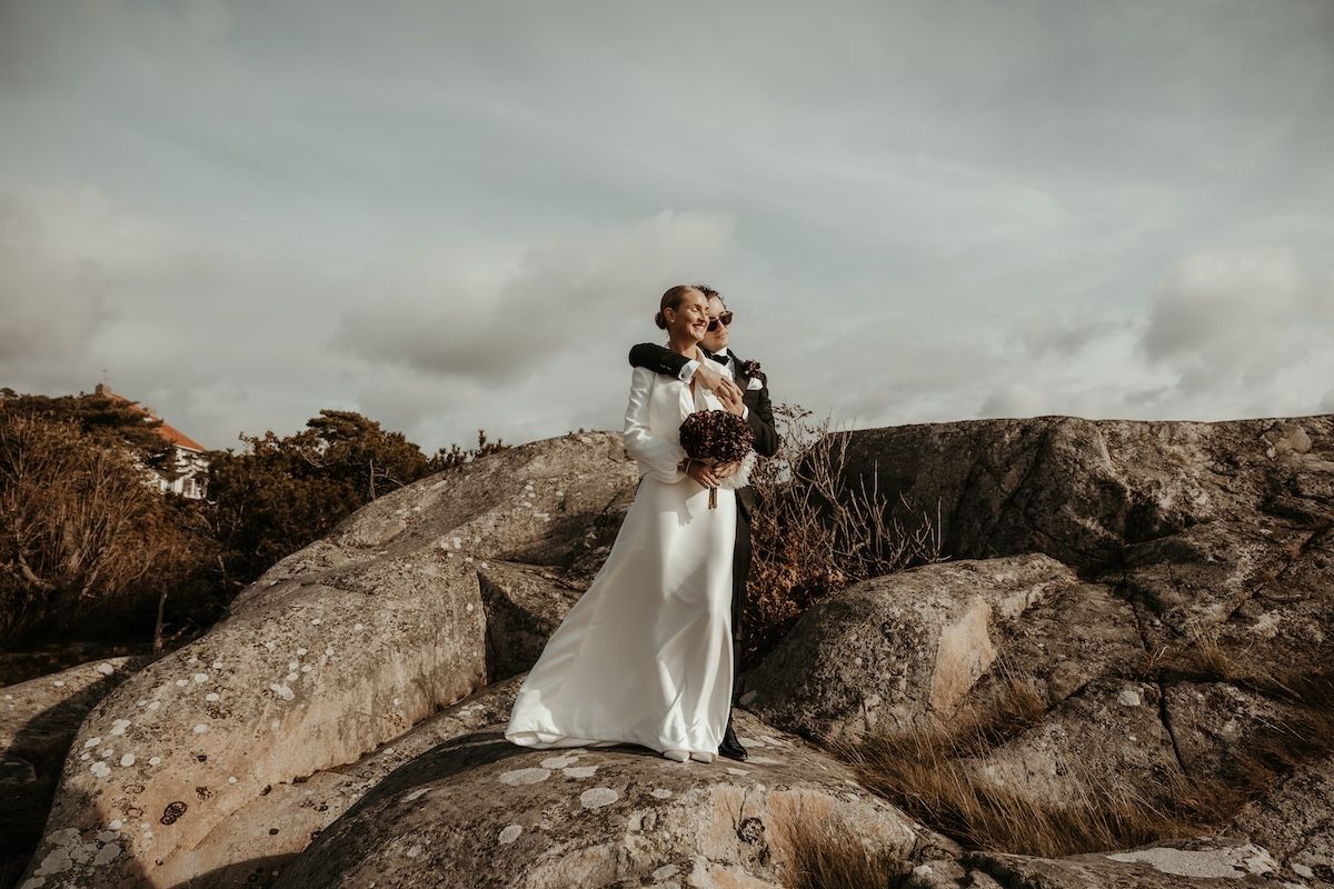 Ett elegant brudpar håller om varandra ståendes på klipporna vid havet under deras västkustbröllop. Foto av Grevholm Photography