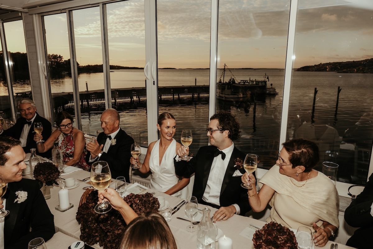 Ett brudpar sitter vid honnörsbordet och håller upp sina glas för att skåla med gästerna. Foto av Grevholm Photography.