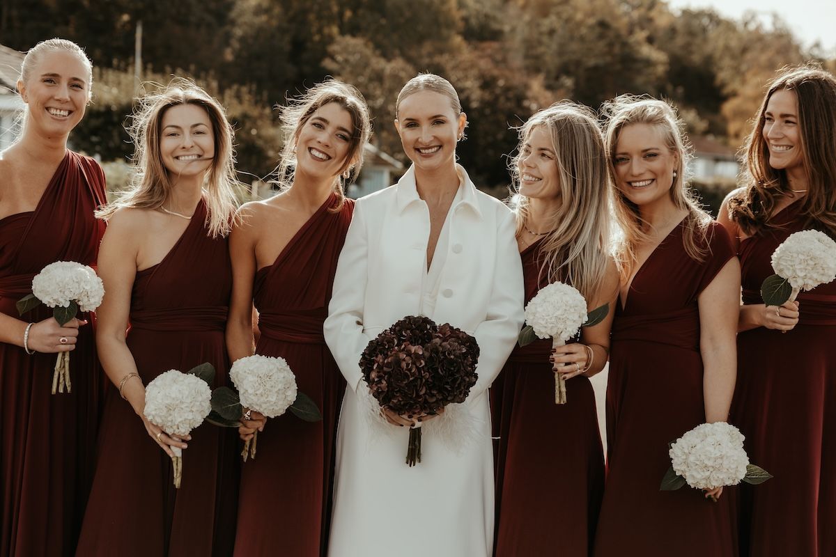 Gruppbild på elegant brud iklädd vitt och hennes brudtärnor iklädda bordeauxfärgade klänningar. Foto av Grevholm Photography