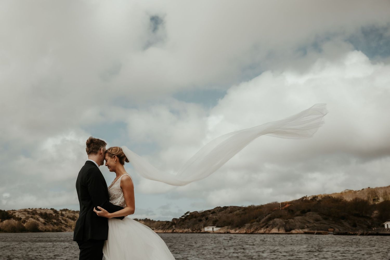 Brudpar står på en brygga i Nösund, Orust och brudslöjan blåser högt i vinden. Fotograf: Grevholm Photography