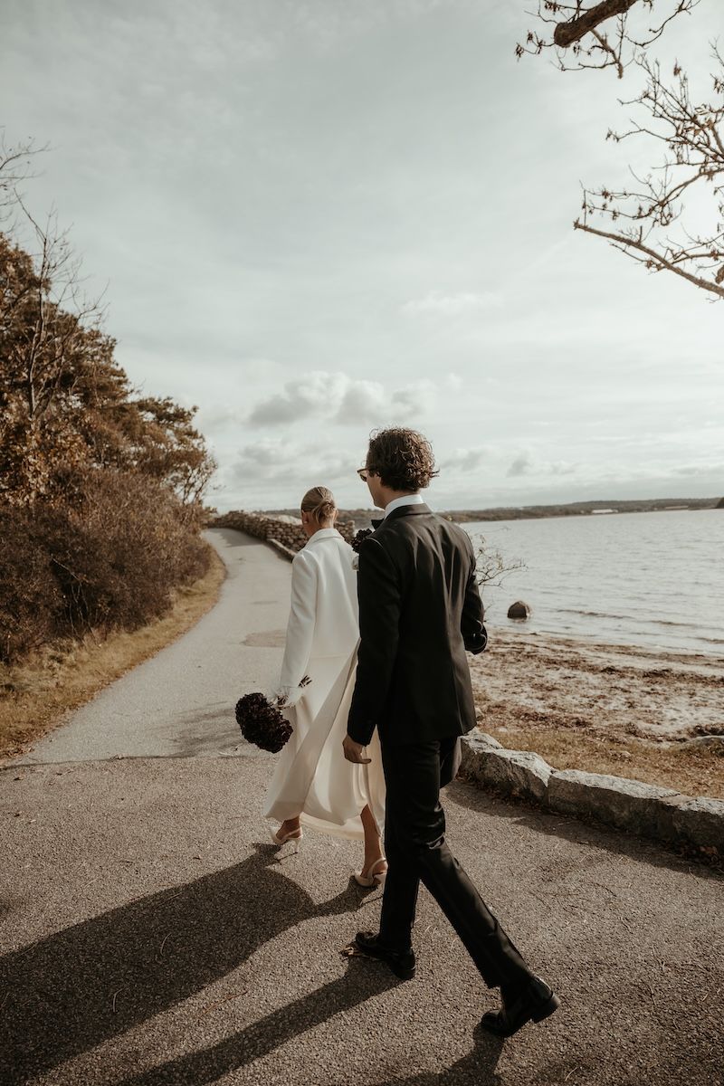 Ett elegant brudpar går hand i hand längs en väg under porträttfotograferingen på deras bröllop på Särö. Foto av Grevholm Photography