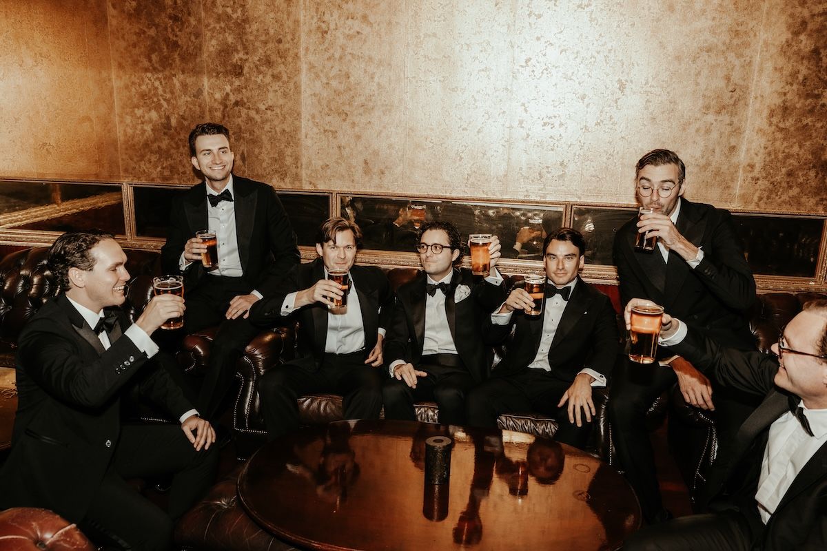 En brudgum och hans marskalker iklädda svarta kostymer sitter i en bar och skålar i öl. Foto av Grevholm Photography