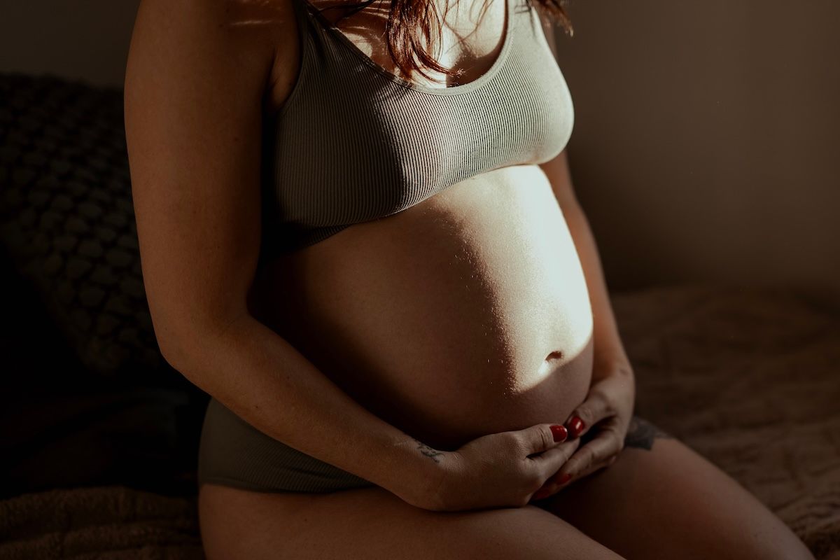 Gravid kvinna sitter på sängkanten iklädd underkläder under en gravidfotografering i hemmet. Foto: Grevholm Photography.