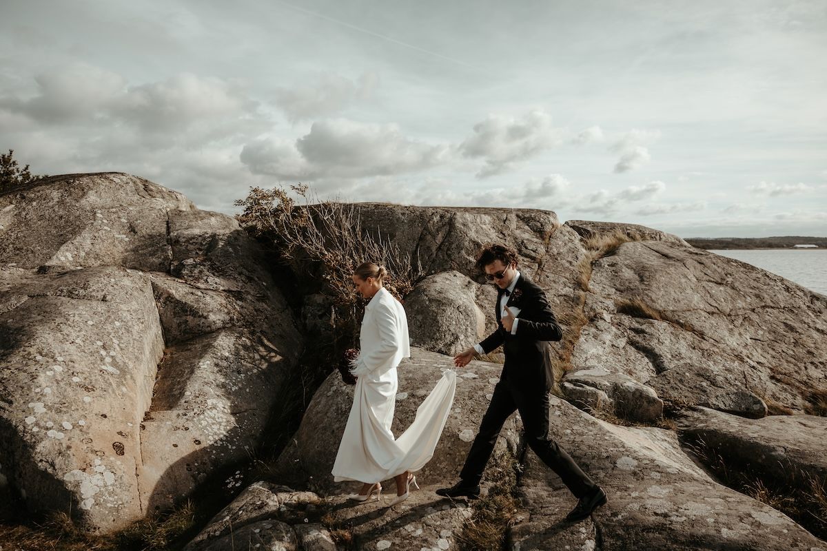 Brudgum håller upp släpet på brudens klänning när de tillsammans går över klipporna vid havet på Särö. Foto av Grevholm Photography