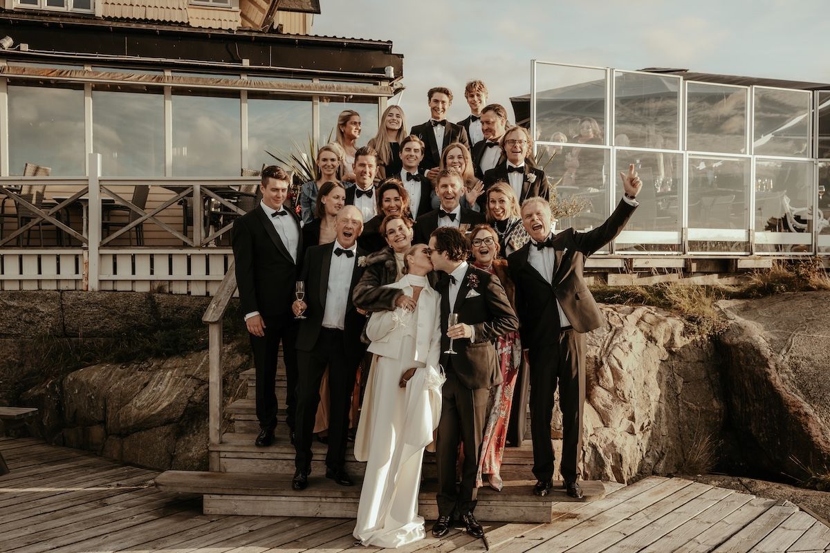 Gruppbild på ett brudpar som kysser varandra och deras familjer som jublar i bakgrunden. Foto av Grevholm Photography