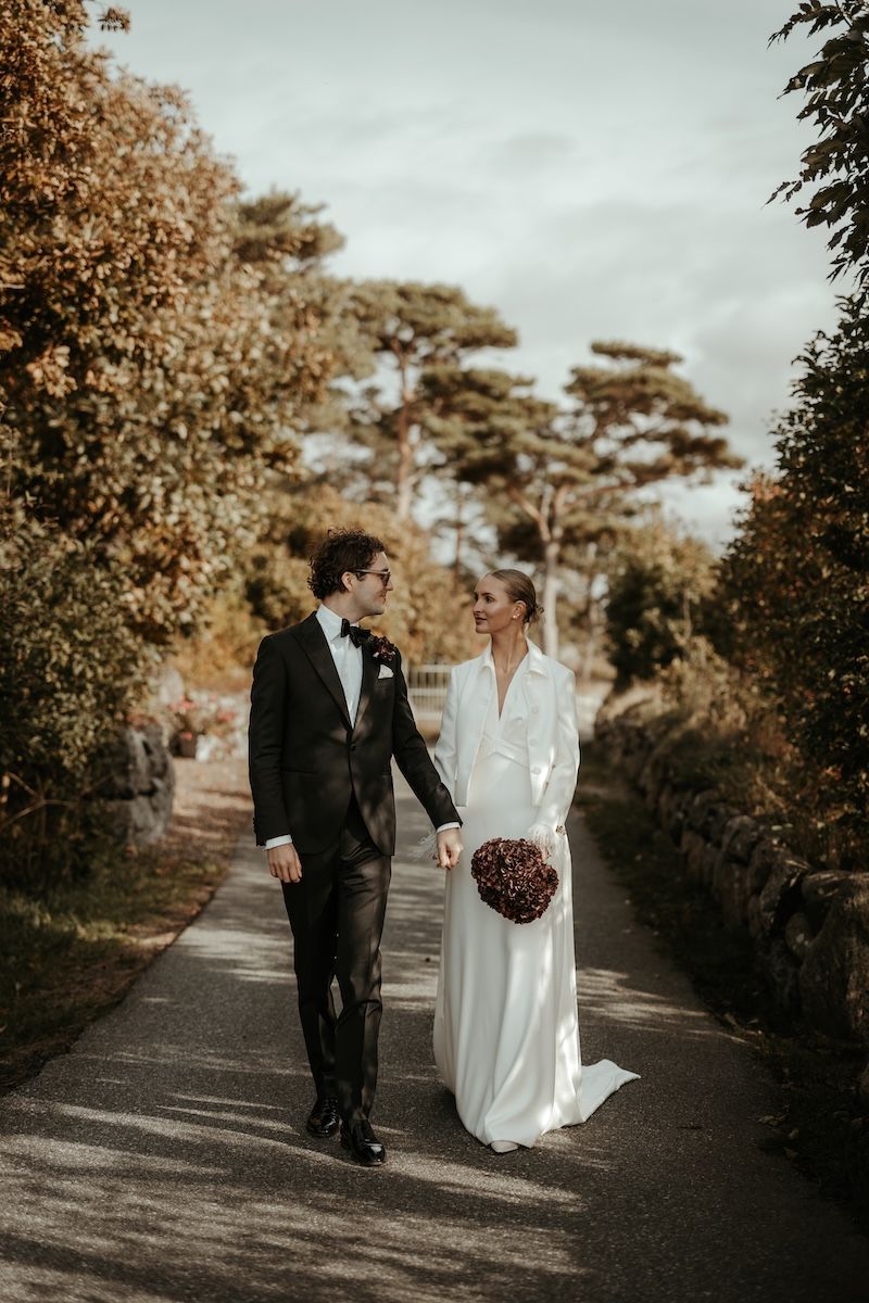 Ett elegant brudpar går hand i hand längs en väg under porträttfotograferingen på deras bröllop på Särö. Foto av Grevholm Photography