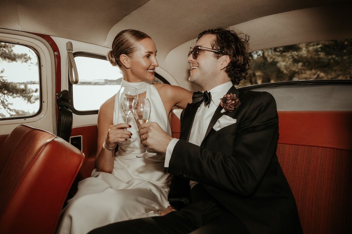 Ett elegant brudpar skålar i champagne sittandes i en gammal bil med röd klädsel. Foto av Grevholm Photography
