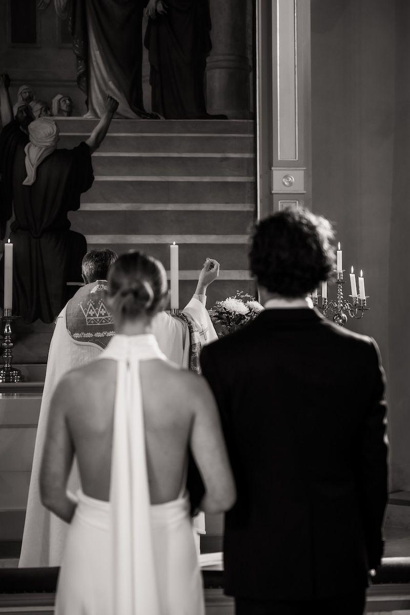 Svartvitt foto av ett brudpar som står vända mot prästen som välsignar ringarna under deras vigselceremoni. Foto av Grevholm Photography
