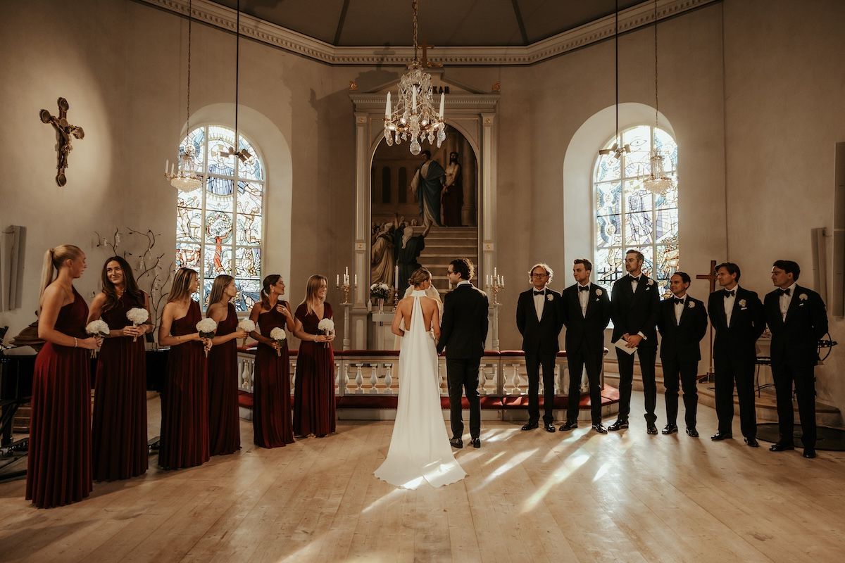 Ett brudpar och deras brudfölje står vid altaret i en kyrka när ljuset strilar in dramatiskt genom ett fönster. Foto av Grevholm Photography