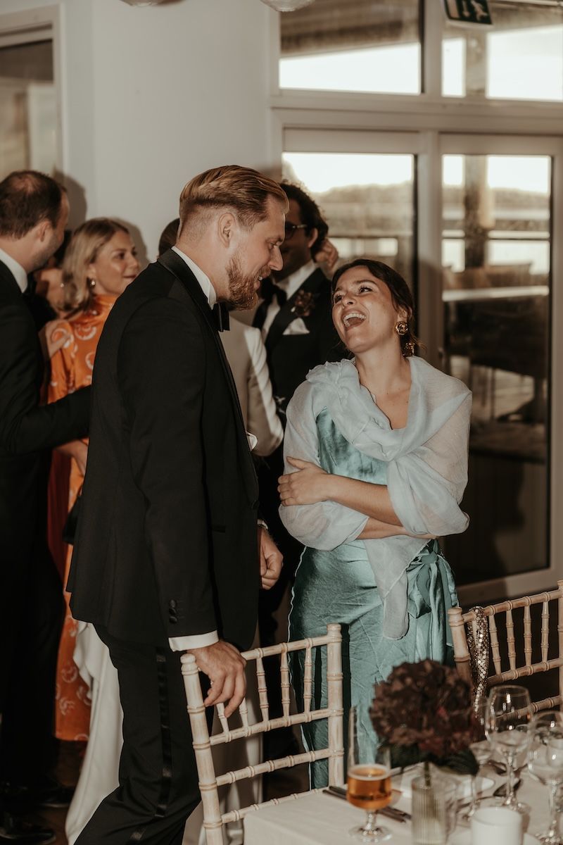 Bröllopsgäster står bakom stolarna i middagslokalen och pratar och skrattar. Foto av Grevholm Photography.