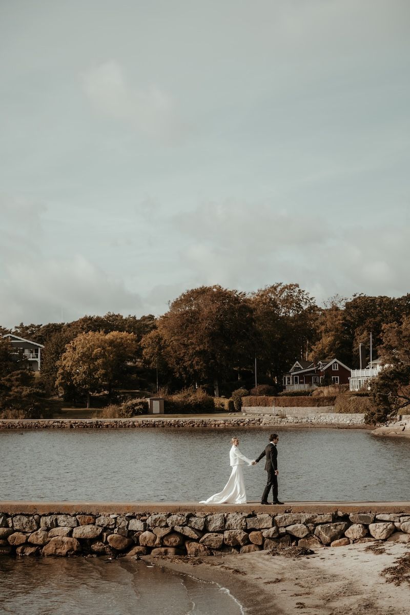 Ett elegant brudpar går hand i hand längs en stenbro i havet under deras bröllop på Särö. Foto av Grevholm Photography