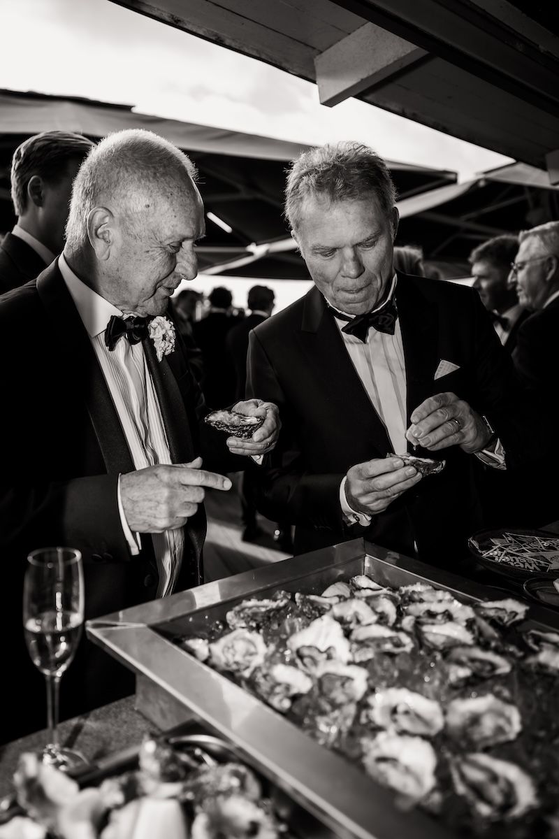Papporna till bruden och brudgummen äter ostron tillsammans under minglet av deras barns bröllop på västkusten. Foto av Grevholm Photography