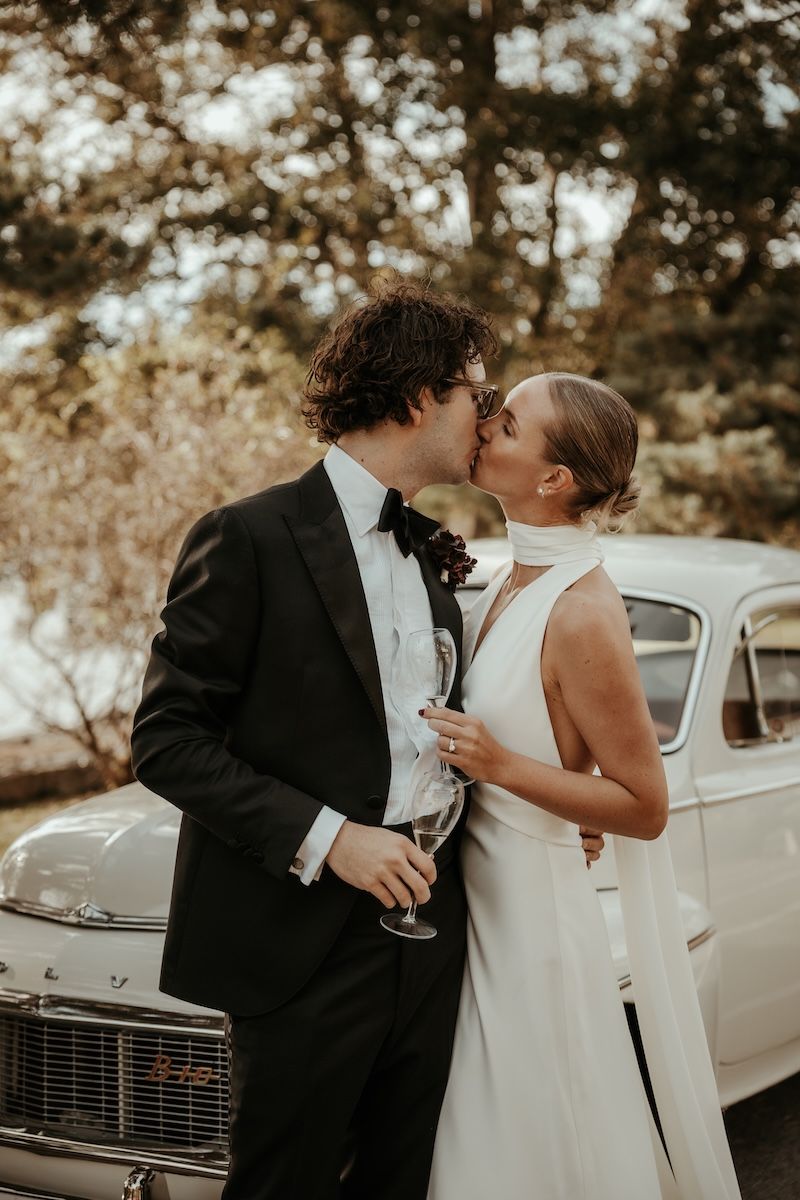 Ett romantiskt brudpar kysser varandra ståendes framför en gammal bil i en varm utomhusmiljö. Foto av Grevholm Photography