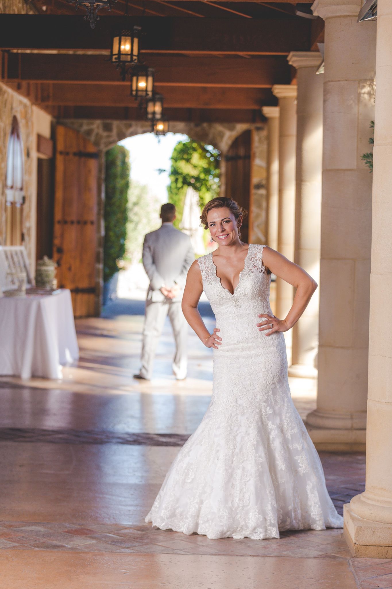 Bride poses in elegant lace wedding dress under covered archway with stone columns.