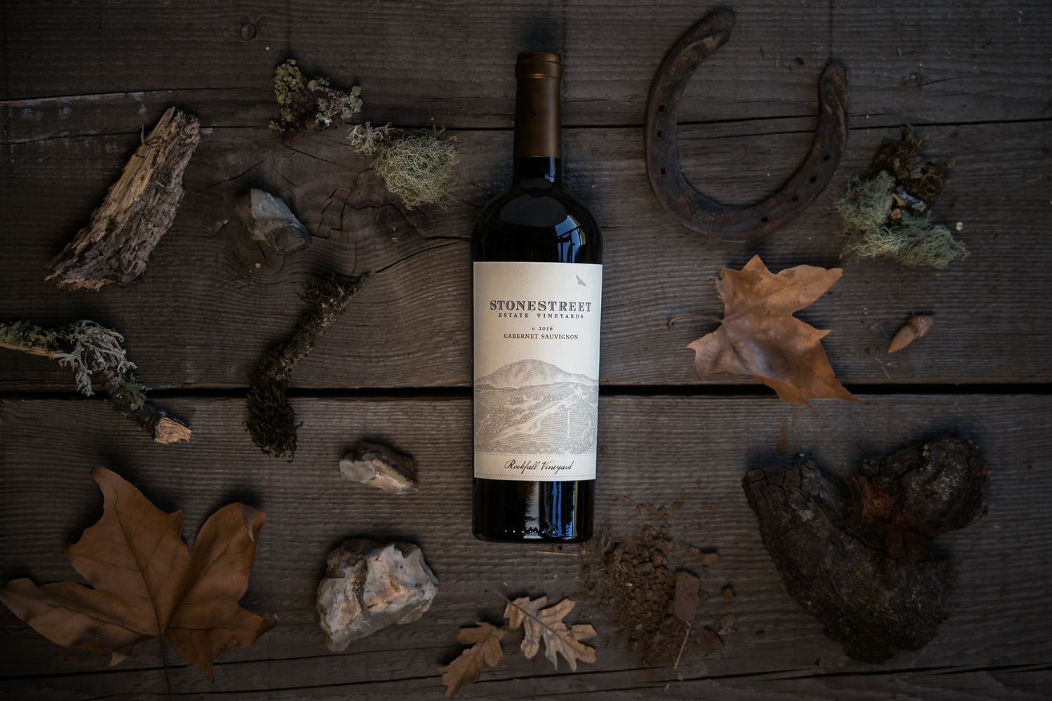 Wine bottle centered among autumn leaves and rustic decor on weathered wood surface.