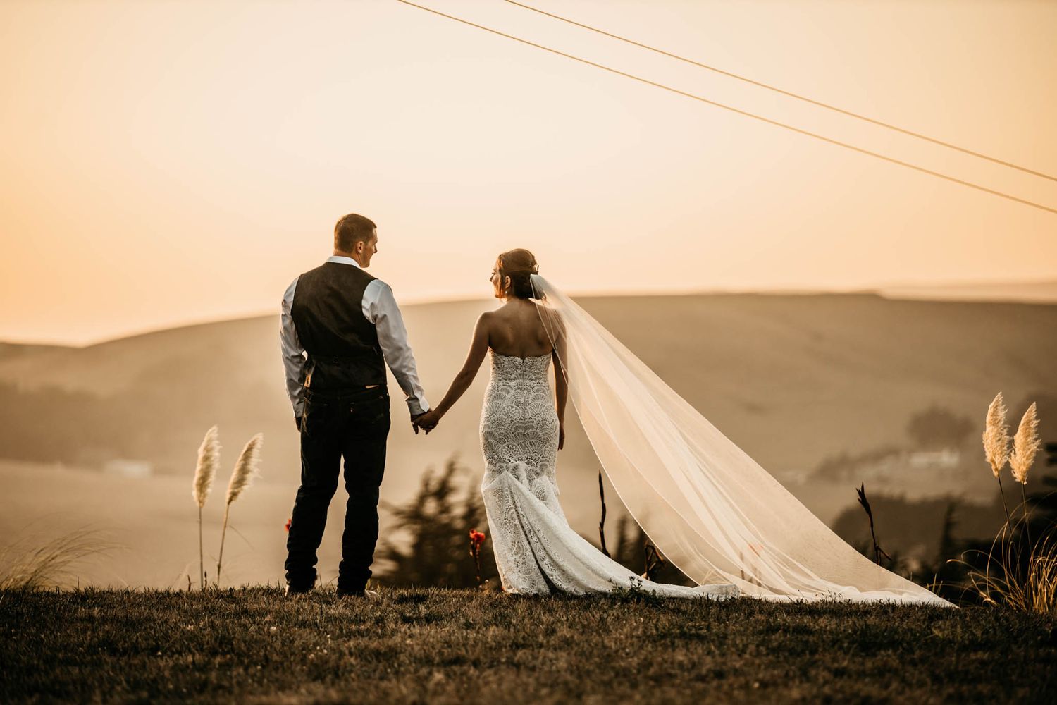 Newlyweds holding hands while walking along a hilltop against a sunset sky.