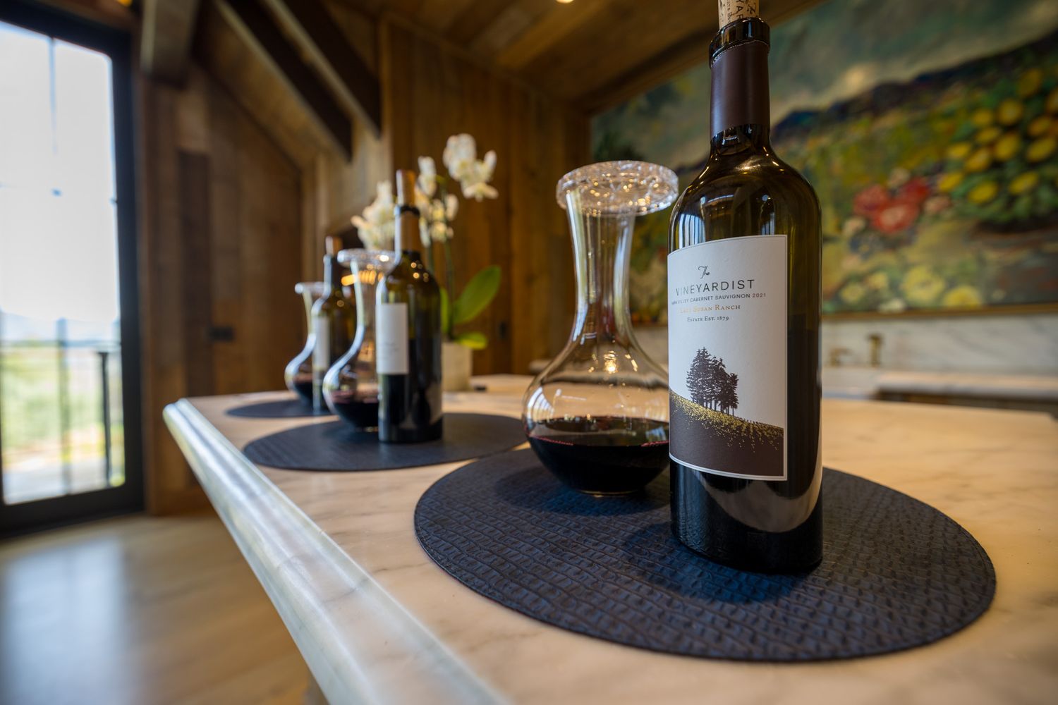 Wine bottles and decanters arranged on elegant black placemats along a white marble counter.