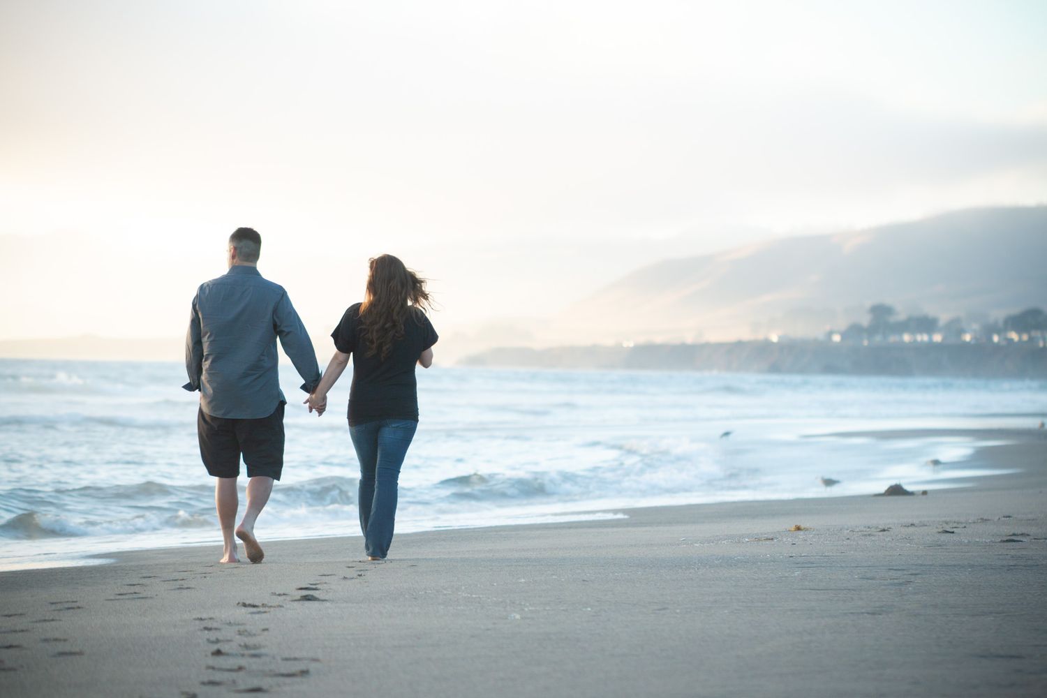 Couple walking hand in hand on empty beach with mountains in distance.